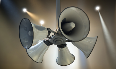 An array of five horn loudspeakers hanging off a pole in a circle facing outwards on a spotlit event background